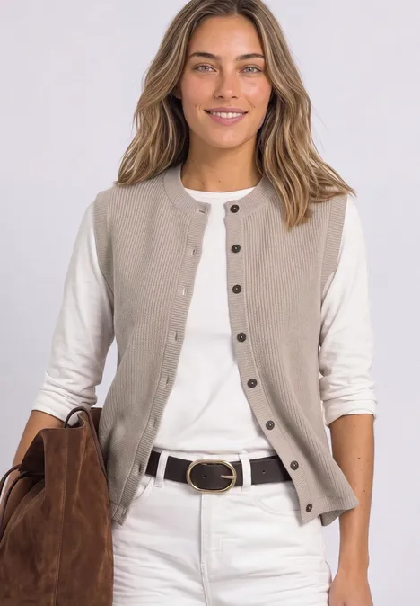 Frau mit beigem Ärmellosen Cardigan, weißem Langarmshirt, weißer Jeans und brauner Tasche lächelt.