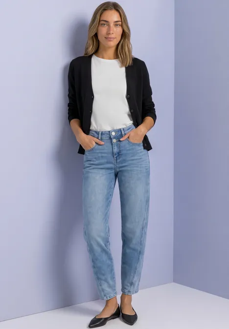 Eine lächelnde Frau im weißen Shirt, schwarzer Strickjacke und blauen Jeans vor lila Wand.