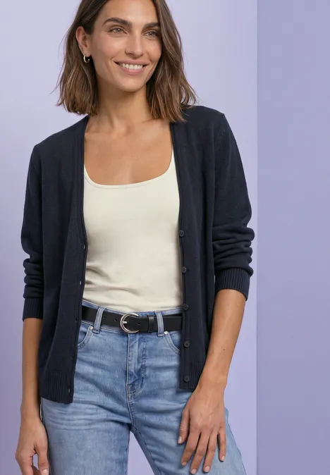 Frau in schwarzer Strickjacke, beigem Top und Jeans vor lila Hintergrund.