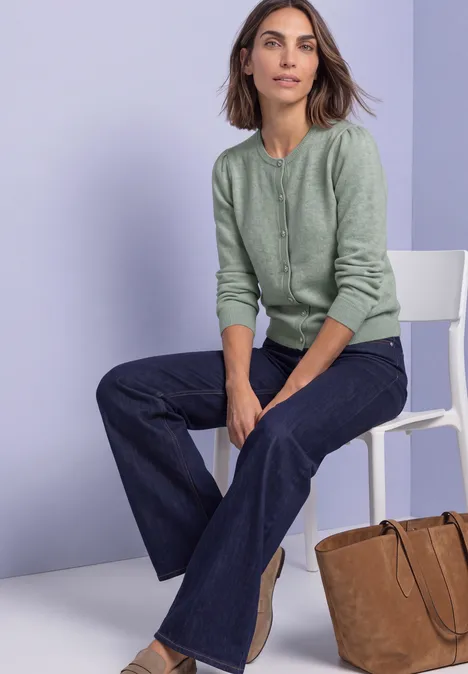 Langarm Cardigan mit Rundhals und Knöpfen eucalyptus green mel. Frau in mintgrünem Cardigan und weiten Jeans sitzt auf weißem Stuhl vor dezentem Pastellhintergrund.
