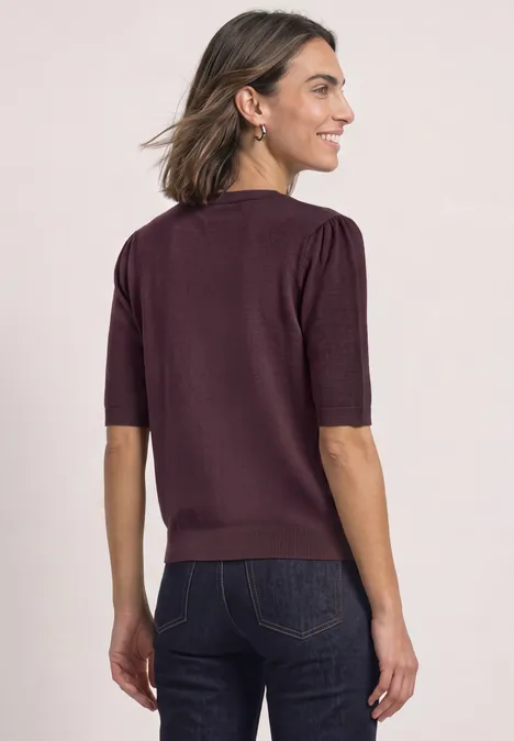 Frau mit bordeauxfarbigem Kurzarmpullover und dunkler Jeans, Rücken zur Kamera, seitlich schauend.