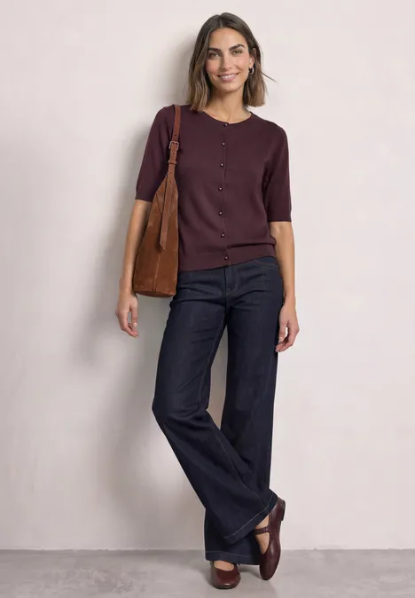 Frau in lila Oberteil und ausgestellten Jeans, trägt Tasche und locker lächelnd.