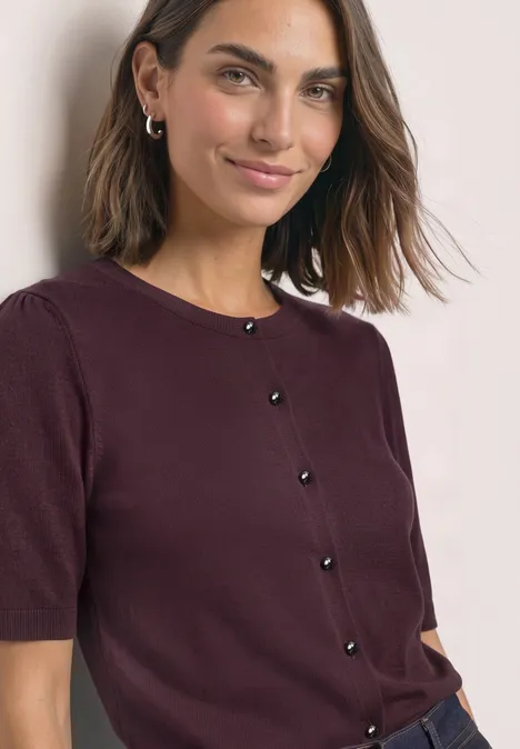 Eine Frau mit schulterlangem Haar trägt eine weinrote Kurzarmstrickjacke mit schwarzen Knöpfen und lächelt.
