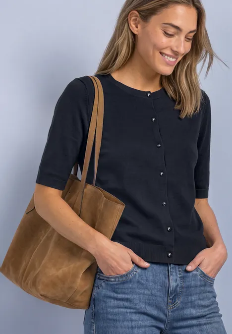 Eine Frau trägt eine schwarze Strickjacke, Jeans und eine braune Wildleder-Tasche und lächelt.