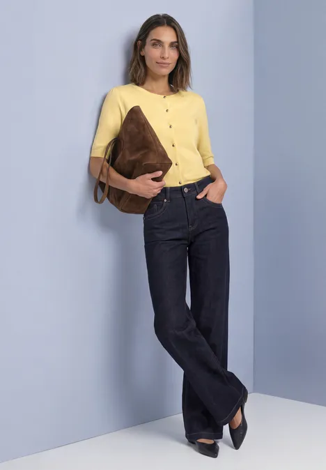 Frau in gelbem Shirt und dunkelblauen Jeans, hält eine braune Tasche und lehnt lässig an einer Wand.