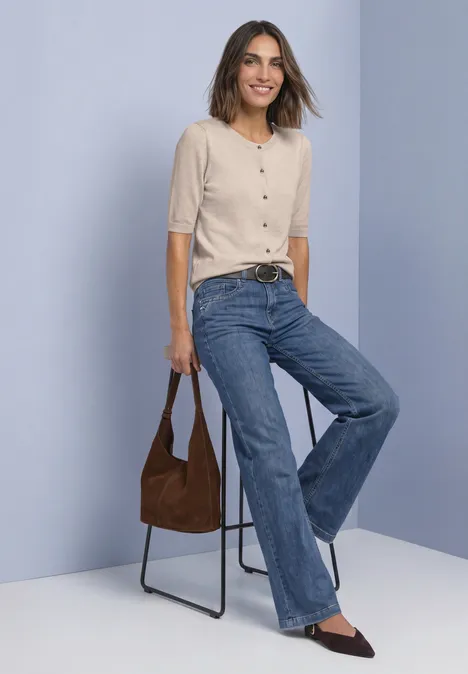 Frau lehnt an Stuhl, lächelt, trägt hellen Pullover, weite Jeans, braune Tasche und spitze Schuhe.