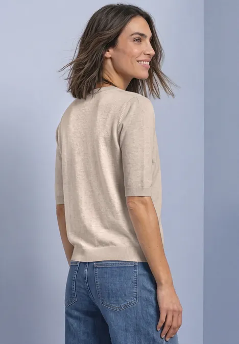 Frau mit halblangem braunem Haar trägt einen beigen Kurzarmpullover und blaue Jeans, Blick über die Schulter.
