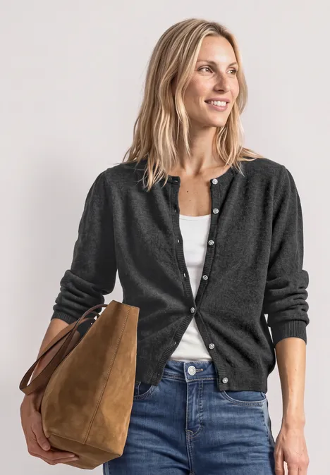 Frau in Jeans, weißem Top und dunklem Cardigan, trägt eine braune Tasche, lächelt entspannt.
