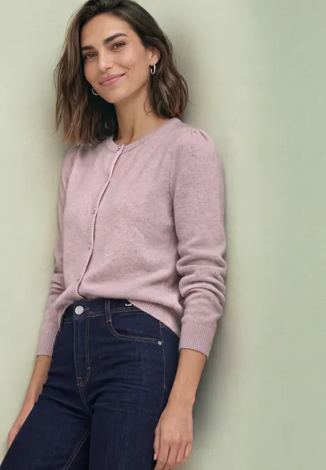Langarm Cardigan mit Rundhals und Knöpfen quiet rose mel. Eine lächelnde Frau in rosa Strickjacke und dunkler Jeans lehnt leicht an einer Wand.