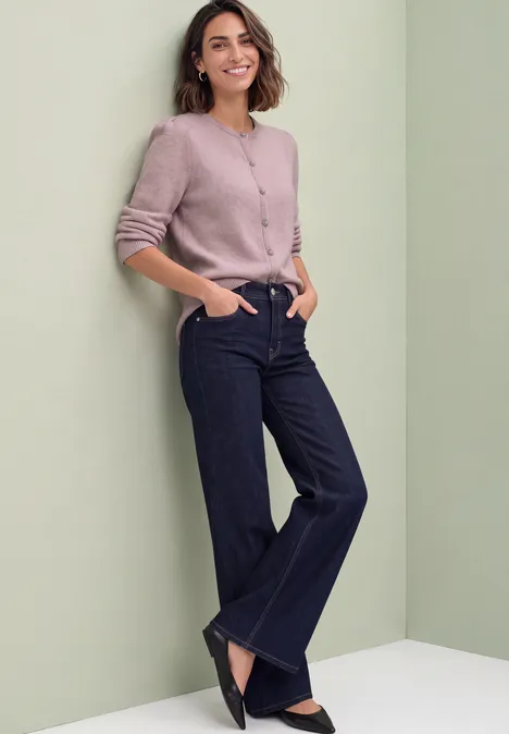 Langarm Cardigan mit Rundhals und Knöpfen quiet rose mel. Eine lächelnde Frau lehnt entspannt an einer grünen Wand, trägt rosa Cardigan und dunkelblaue Jeans.