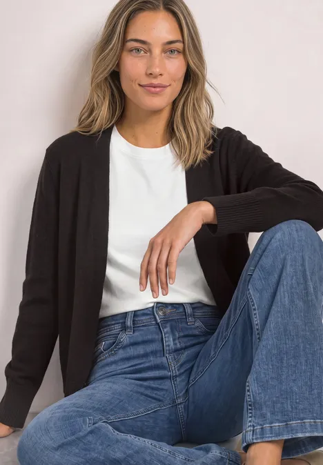 Frau sitzt, trägt weiße T-Shirt, schwarze Strickjacke und blaue Jeans, posiert vor hellem Hintergrund.