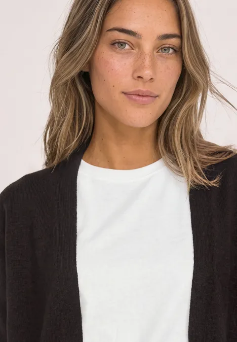 Frau mit schwarzem Cardigan und weißem T-Shirt, Schulterporträt vor hellem Hintergrund, leichtes Lächeln.
