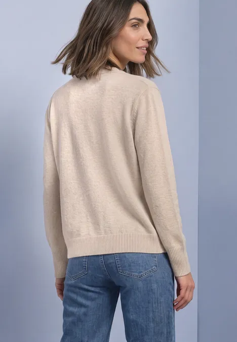 Une femme de dos porte un pull beige et un jean bleu devant un fond uni.