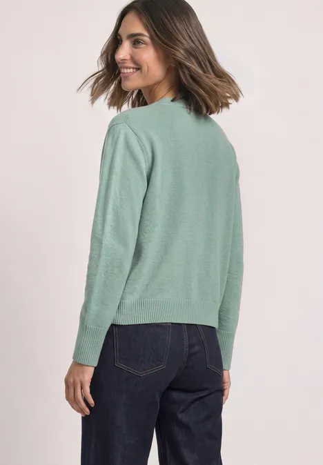 Eine Frau trägt einen hellgrünen Pullover und dunkle Jeans, blickt lächelnd über die Schulter.