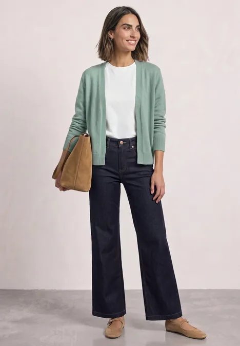 Frau in weißem Shirt, mintgrüner Strickjacke, dunklen Jeans, beigen Slippern und Tasche.