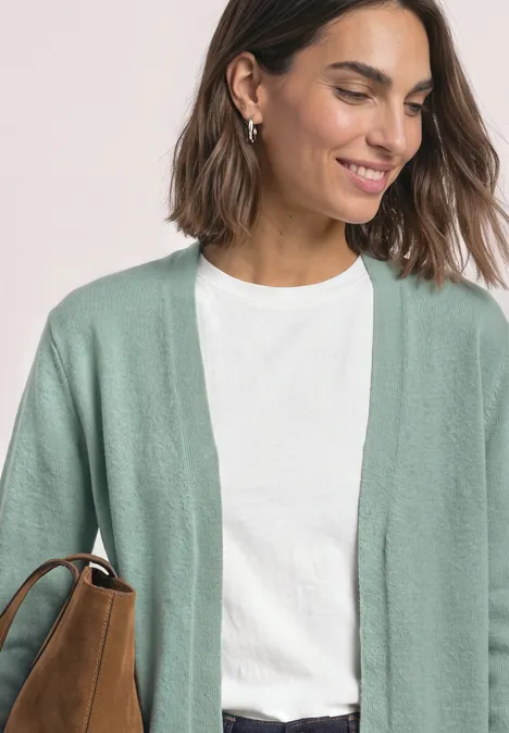 Frau mit weißem T-Shirt und mintgrünem Cardigan, lächelnd mit brauner Tasche über der Schulter.