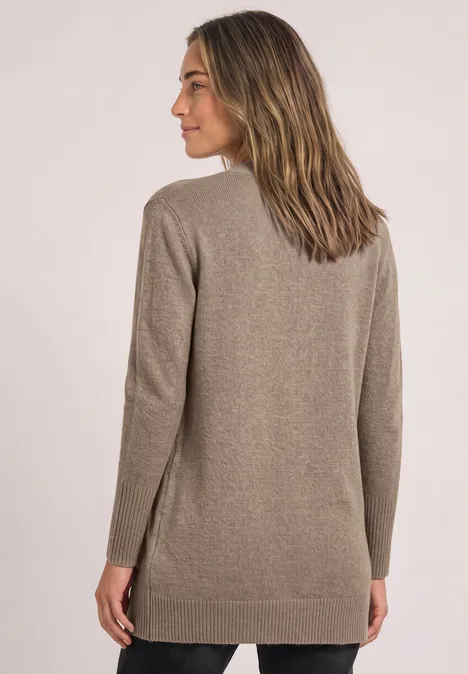 Frau in taupefarbenem, langem Strickcardigan, steht halbseitlich vor hellem Hintergrund und blickt nach links.