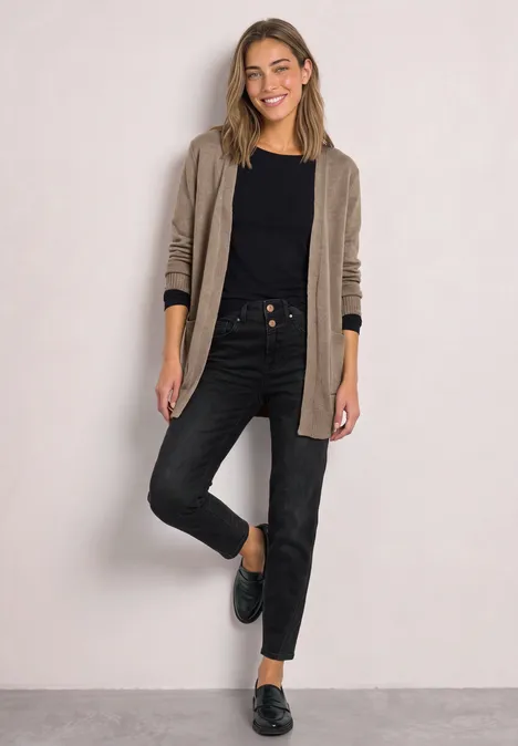 Junge Frau in beigem Cardigan, schwarzem Shirt, dunklen Jeans und schwarzen Slippern vor hellem Hintergrund.