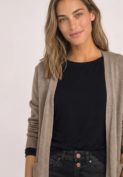 Eine lächelnde Frau mit braunen Haaren trägt ein schwarzes Shirt und eine beige Strickjacke vor hellem Hintergrund.