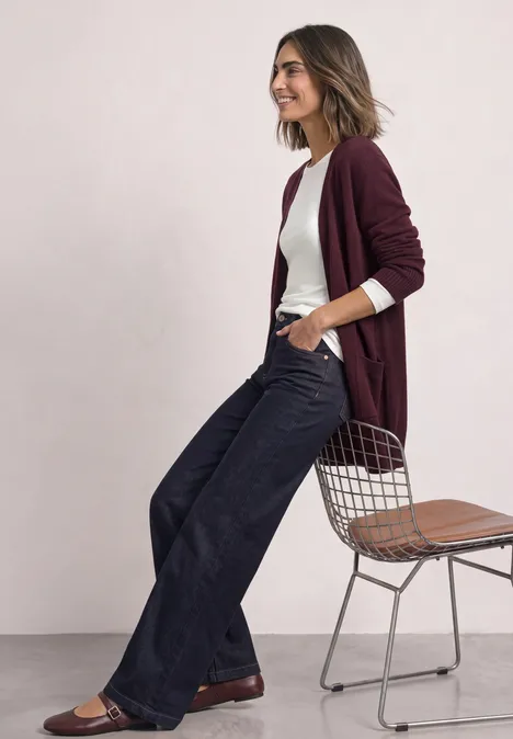 Femme en cardigan bordeaux, t-shirt blanc et jeans évasés, appuyée et souriante contre une chaise.