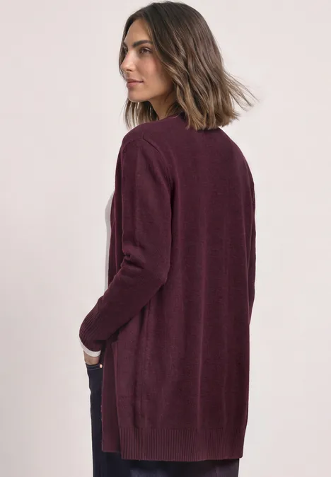 Femme de profil portant un long cardigan bordeaux, regard tourné sur le côté.