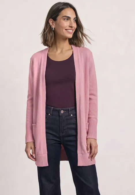 Frau trägt rosa Strickjacke über dunklem Top und Jeans, lächelt selbstbewusst.
