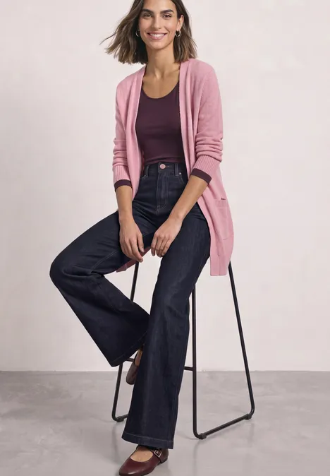 Frau sitzt lässig auf einem Hocker, trägt rosa Cardigan, lila Top und dunkle Schlagjeans.