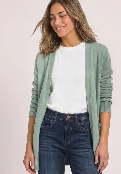 Frau mit weißem T-Shirt, blauen Jeans und grüner Strickjacke lächelnd