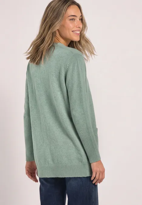 Eine lächelnde Frau mit blond-braunen Haaren trägt einen grünen Pullover und blickt über die Schulter.