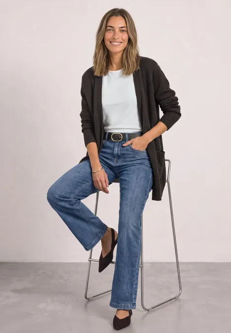 Femme assise sur un tabouret, portant un t-shirt blanc, un cardigan noir et un jean bleu, souriante.