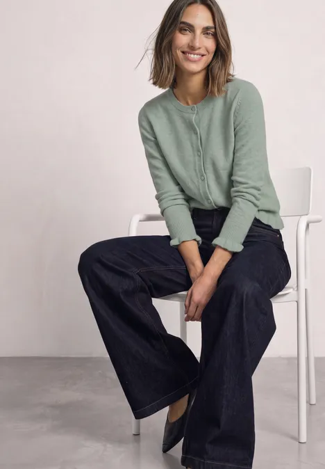 Cardigan mit Rundhals und Rüschendetails eucalyptus green mel. Frau sitzt auf weißem Stuhl, trägt grünen Cardigan und weite dunkle Jeans, lächelt entspannt.