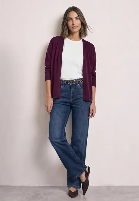 Kardigan z rozpinanym dekoltem w jednolitym kolorze berry maroon Kobieta pozuje na neutralnym tle, ma na sobie biały t-shirt, bordowy kardigan, niebieskie dżinsy i mokasyny.