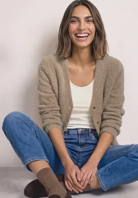 Vrouw zit lachend in vest, top, spijkerbroek en sokken tegen neutrale achtergrond.