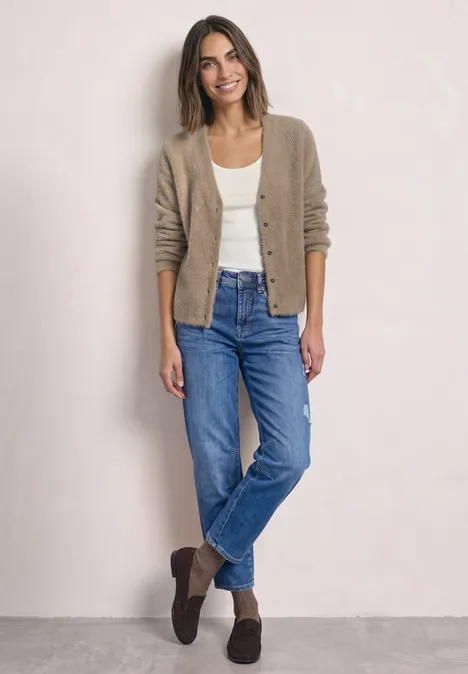 Vrouw in spijkerbroek, wit shirt en beige vest, ontspannen poserend voor een lichte achtergrond.