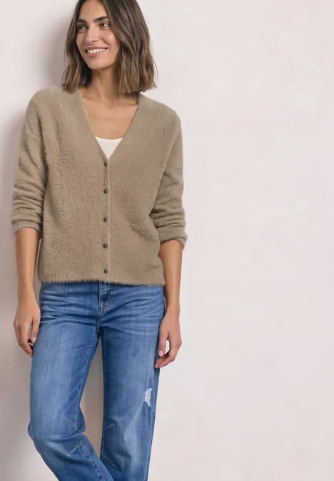 Een vrouw draagt een beige vest en blauwe spijkerbroek, staand glimlachend voor een lichte achtergrond.