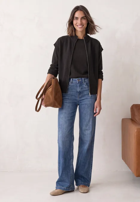 Vest met afhangende schouders, coltrui en ritssluiting black coffee mel. Frau in schwarzer Jacke, schwarzem Top und blauen Jeans hält eine braune Tasche vor hellem Hintergrund.