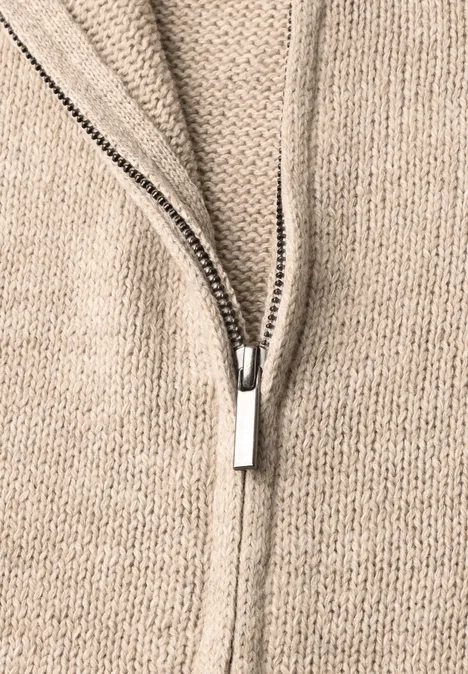 Close-up van een beige gebreide trui met een deels open zilverkleurige ritssluiting