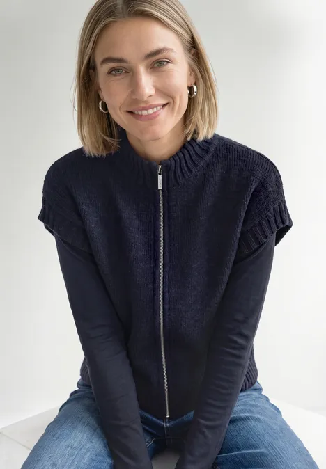 Vest met afhangende schouders, coltrui en ritssluiting indigo night mel. Lächelnde blonde Frau sitzt in Jeans und blauem Zip-Pullover und blickt entspannt in die Kamera.