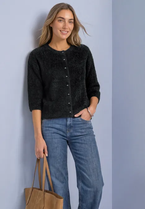 Frau in schwarzer Strickjacke und Jeans, hält Tasche und lächelt vor schlichtem Hintergrund.