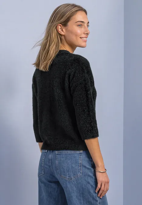 Eine junge Frau in Jeans und einem schwarzen, flauschigen Pullover blickt lächelnd seitlich über ihre Schulter.
