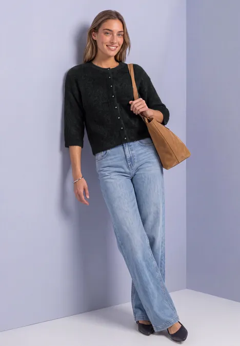Eine lächelnde junge Frau lehnt an einer hellvioletten Wand, trägt schwarzen Cardigan, Jeans und Tasche.