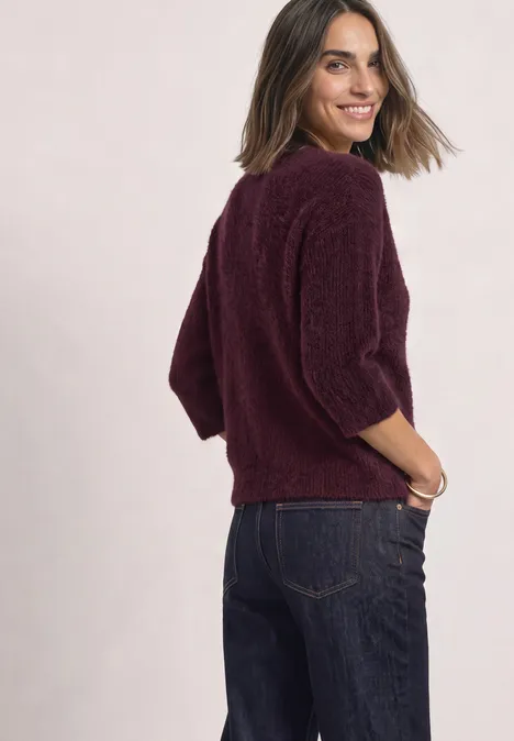 Eine lächelnde Frau in einer bordeauxroten Strickjacke und Jeans, schaut über die Schulter.