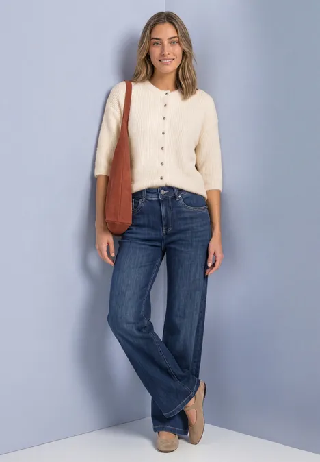 Eine lächelnde Frau in blauen Jeans und cremefarbenem Cardigan mit brauner Tasche vor hellem Hintergrund.