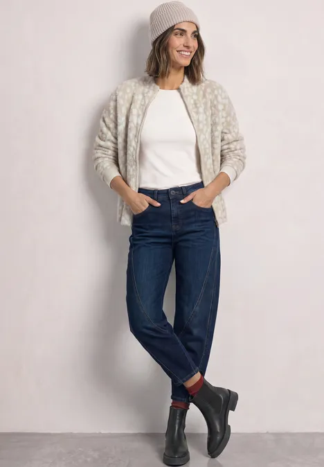 Frau in Jeans, weißem T-Shirt, Cardigan, Beanie und Boots, steht vor hellem Hintergrund und lächelt.