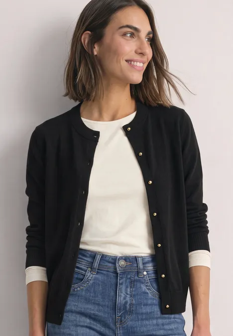 Cardigan à col rond et patte de boutonnage Black Lächelnde Frau in schwarzer Strickjacke, weißem Shirt und Jeans vor neutralem Hintergrund.
