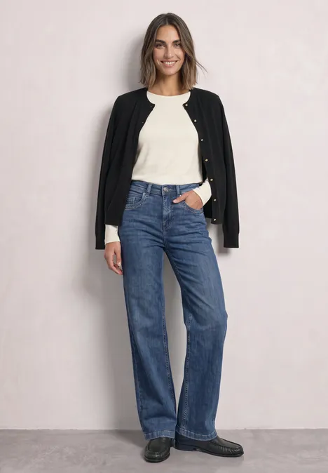 Cardigan à col rond et patte de boutonnage Black Eine Frau vor hellem Hintergrund trägt weiße Bluse, schwarze Jacke über den Schultern und gerade blaue Jeans.
