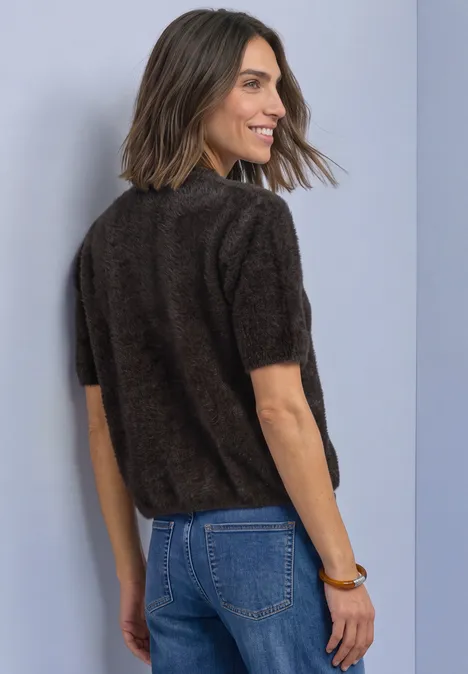 Een glimlachende vrouw in een zacht bruin kort mouwtrui en blauwe jeans kijkt over haar schouder.