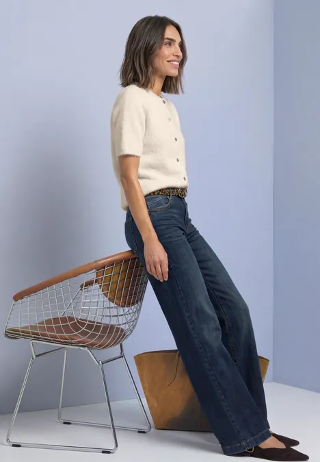 Femme en t-shirt beige et jean large, debout près d'une chaise en fil métallique et d'un bloc de bois sur fond clair.