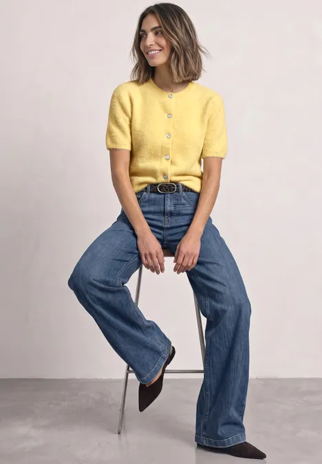 Kurzarm Cardigan mit Rundhals und Knöpfen sunbeam yellow Frau trägt gelbes Oberteil und blaue Jeans und sitzt lässig auf einem Hocker.