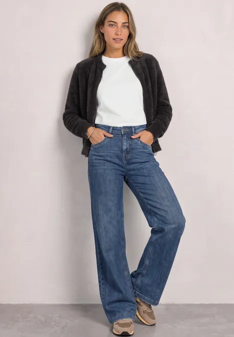 Cardigan en laine fine avec fermeture éclair black coffee Frau in weißem T-Shirt, schwarzer Jacke und weitem Jeans lehnt lässig vor einer hellen Wand.